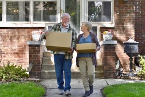Older couple carrying moving boxes outside their home while preparing to downsize for retirement.