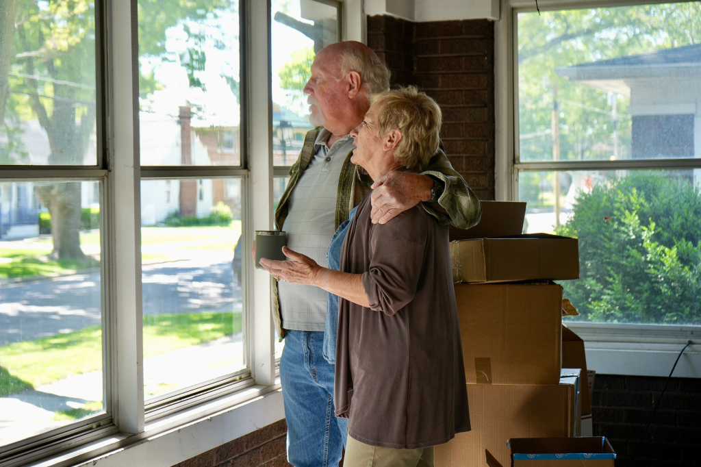 Older couple standing together by a window in their home, with moving boxes stacked behind them.
